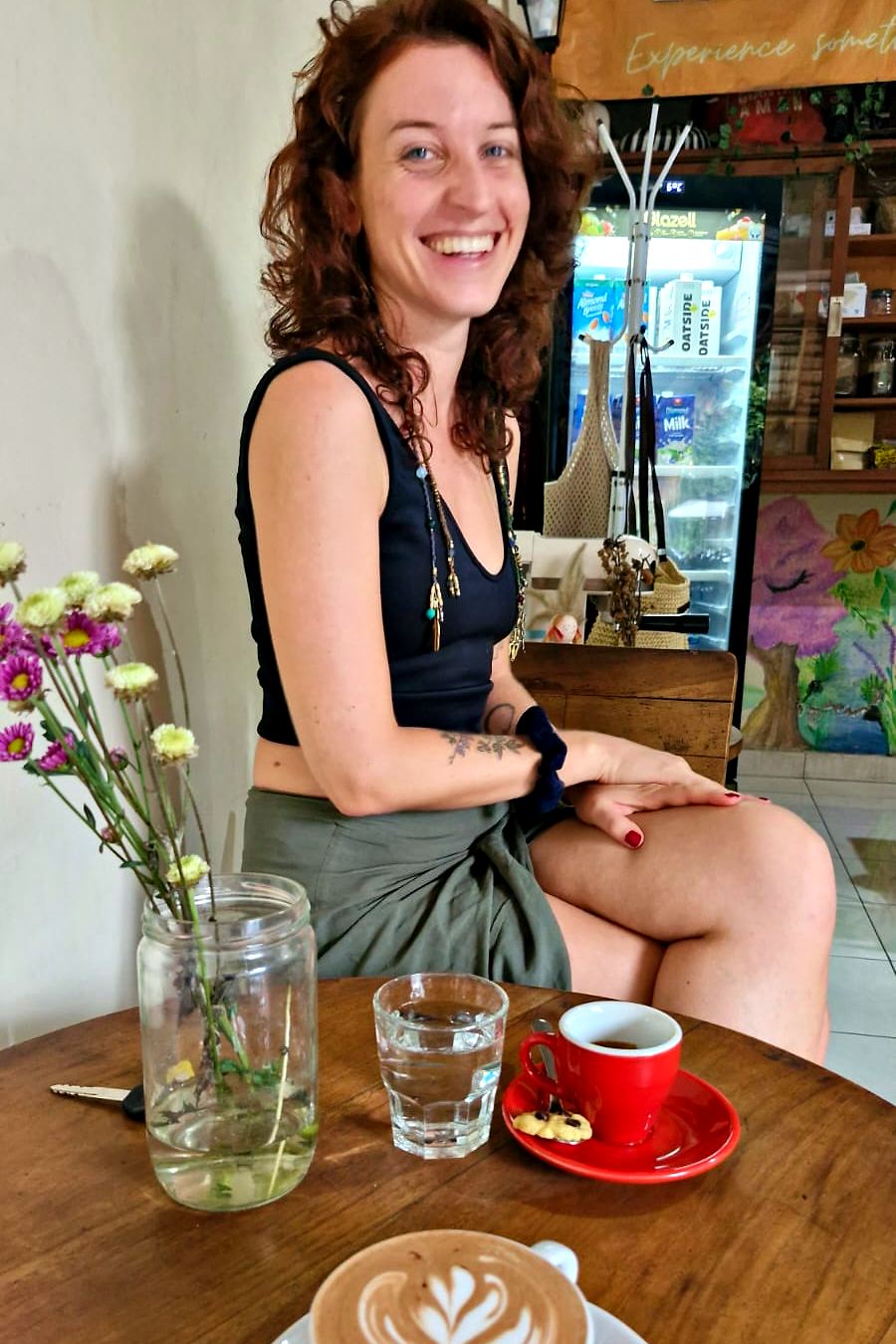 Barbara lacht an einem Cafétisch mit Kaffee und einem Glas Wildblumen.