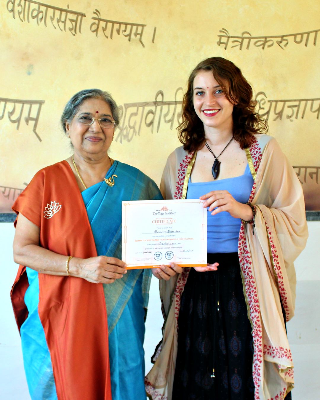 Barbara holding her certificate beside a teacher at The Yoga Institute, in front of a wall painted with Sanskrit verses.