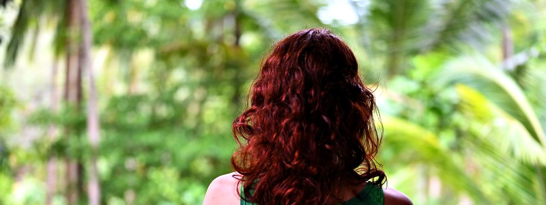 A woman with long red hair seen from behind, looking out over a tropical garden.
