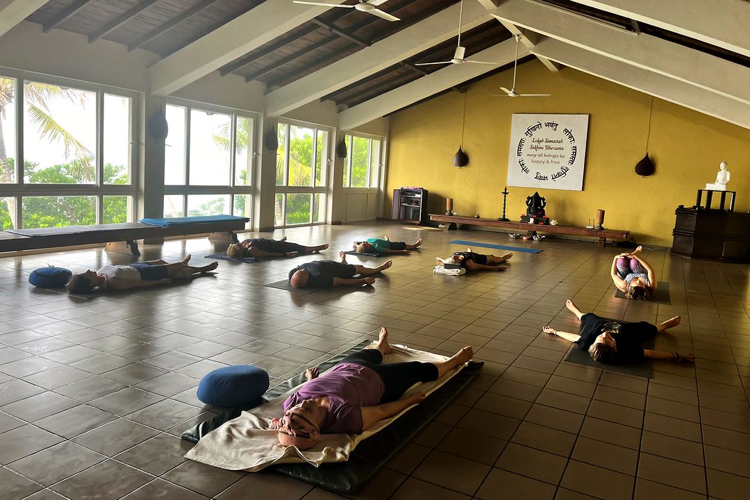 Eine Yogahalle mit Deckenventilatoren und hohen Fenstern, Schülerinnen und Schüler ruhen auf Matten mit Bolstern.